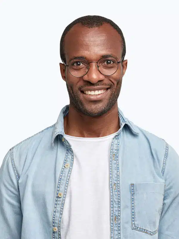 A man with short hair and glasses is smiling at the camera. He is wearing a light denim shirt over a white tee against a plain background.