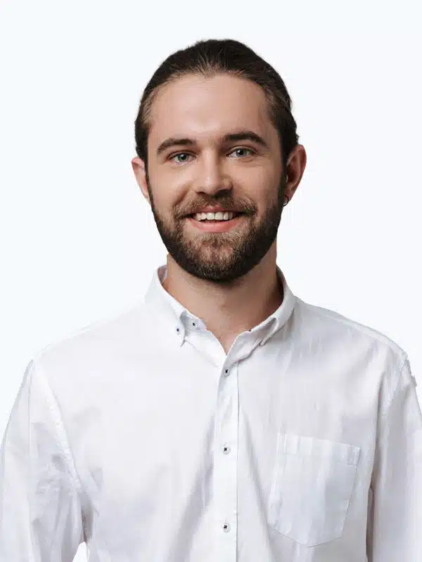 Man with a beard, smiling, wearing a white button-up shirt, and standing against a plain white background.