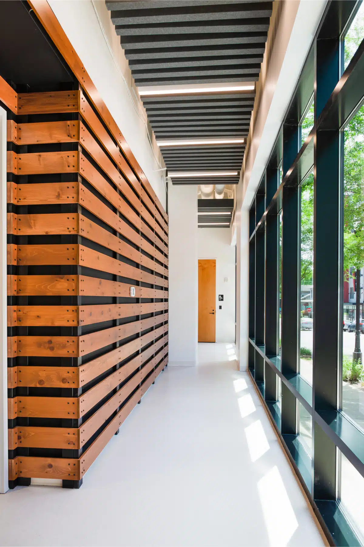 A modern corridor with wooden slat wall panels, large windows on the right, and a wooden door at the end. The ceiling features black acoustic baffles.