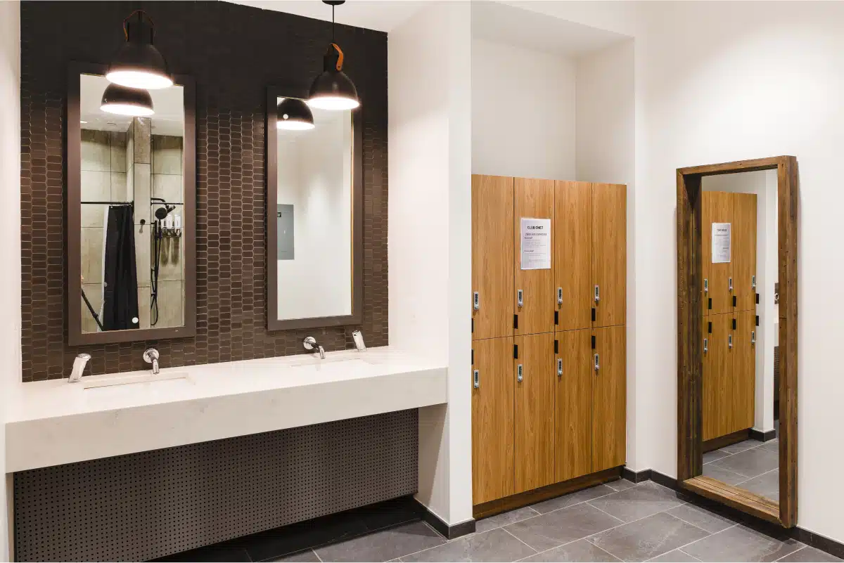 A locker room featuring wooden lockers, a large mirror, a vanity with two sinks, and two mirrors under pendant lights.