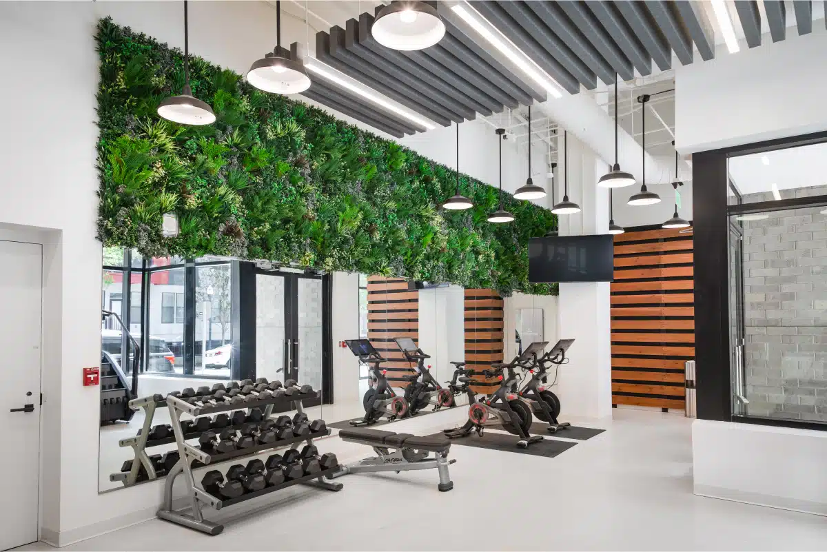 A modern gym with a row of dumbbells, three stationary bikes, a bench, large mirrors, and a wall covered in vertical greenery. The space has high ceilings and pendant lights.