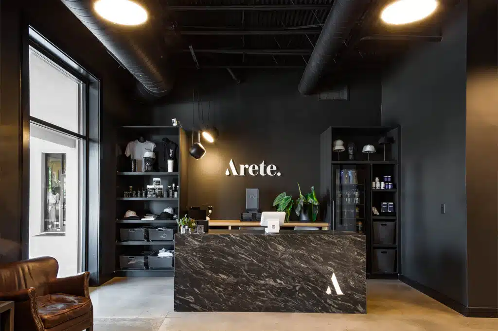 A modern retail store interior with dark walls, a black marble counter, shelves displaying merchandise, and the word "Arete" on the wall behind the counter.