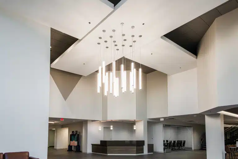 A modern lobby with angular ceilings, a cluster of vertical pendant lights, a central reception desk, and seating area in the background. The area features minimalist design and neutral colors.