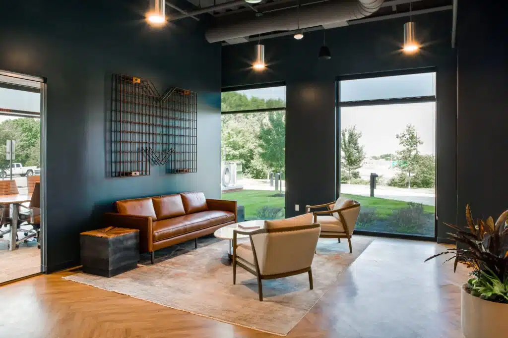 A modern office lounge features a brown leather sofa, two beige armchairs, and a round coffee table on a textured rug, with large windows overlooking a green outdoor area.