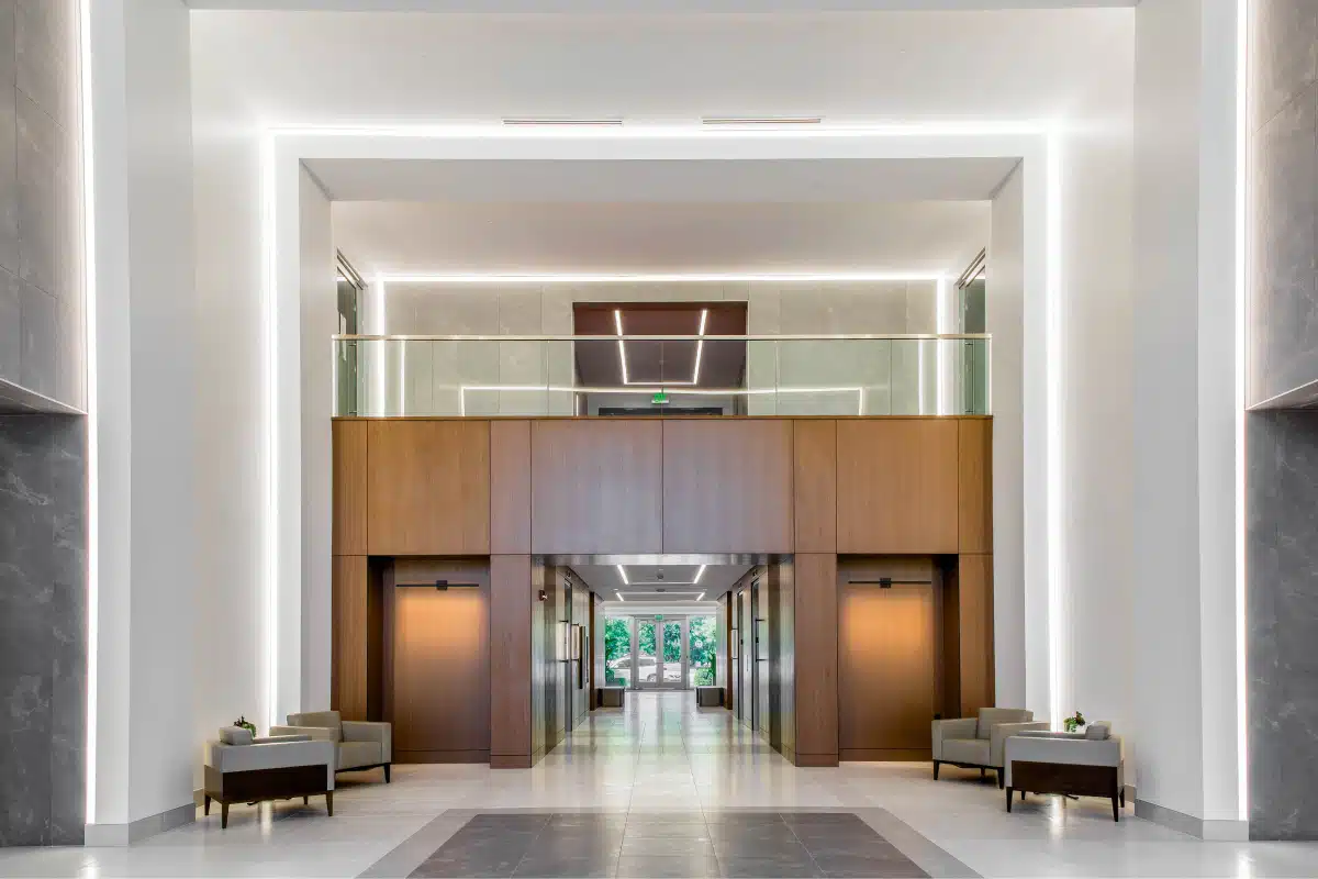 Modern lobby with high ceilings, illuminated coves, and a wooden-paneled second-floor balcony. The area features minimalist furniture, including chairs and tables with small plants.