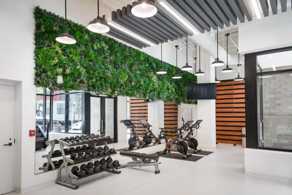 A modern gym with dumbbells, exercise bikes, and a bench. The walls feature a greenery installation and wood paneling. Fluorescent and pendant lights are hanging from the ceiling.