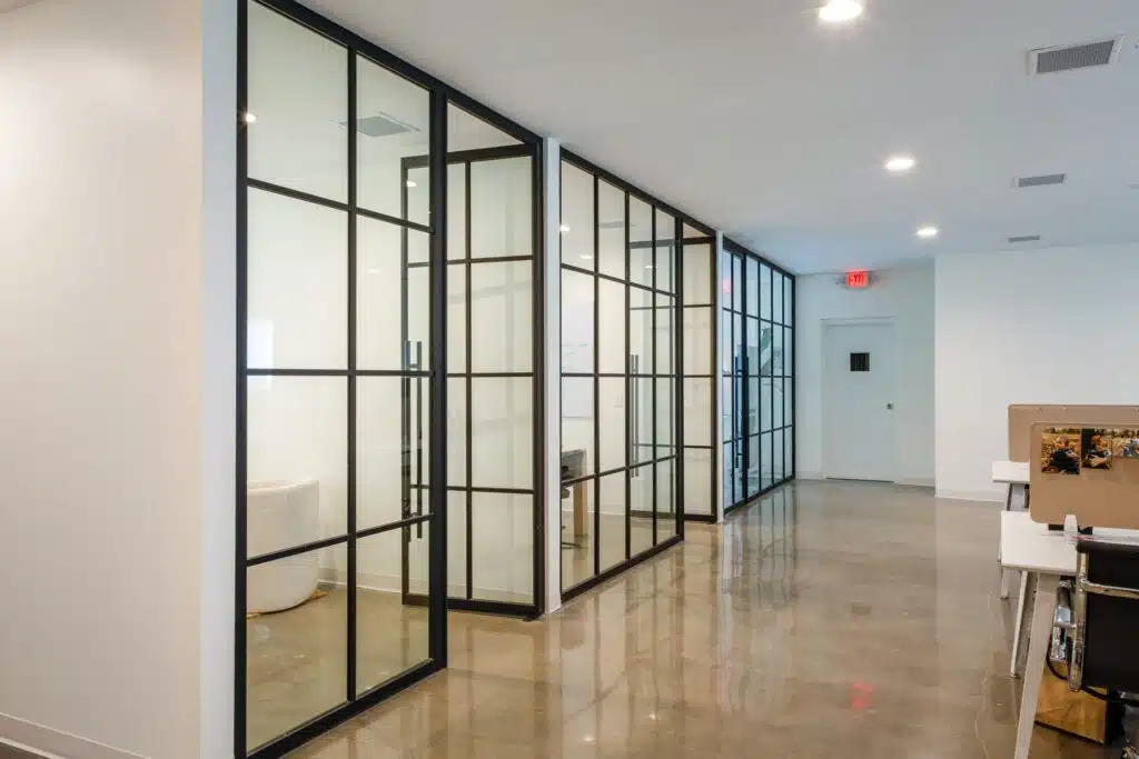 A modern office hallway with glass-paneled doors on the left, concrete flooring, and white walls. At the right end, a partially visible desk and office chair can be seen. A red exit sign is on the ceiling.