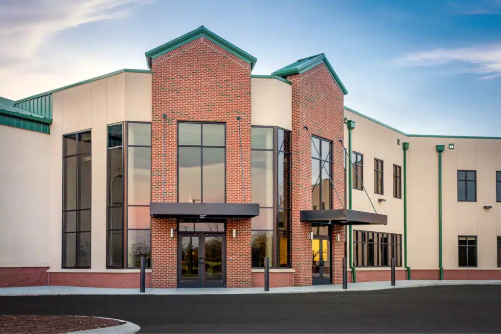 Modern building facade with large glass windows, red brick accents, and a peaked roof under a partly cloudy sky.