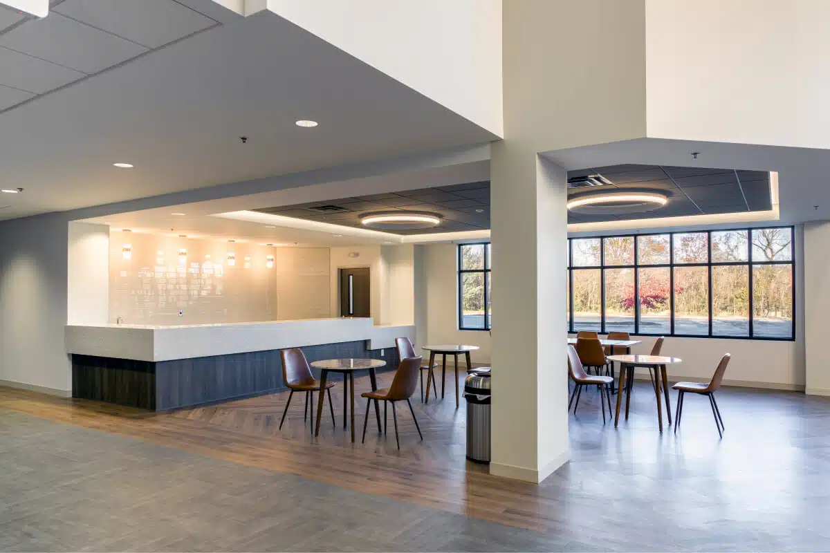 A modern dining area with round tables, wooden chairs, and large windows. The space features a counter with barstools and overhead pendant lighting.