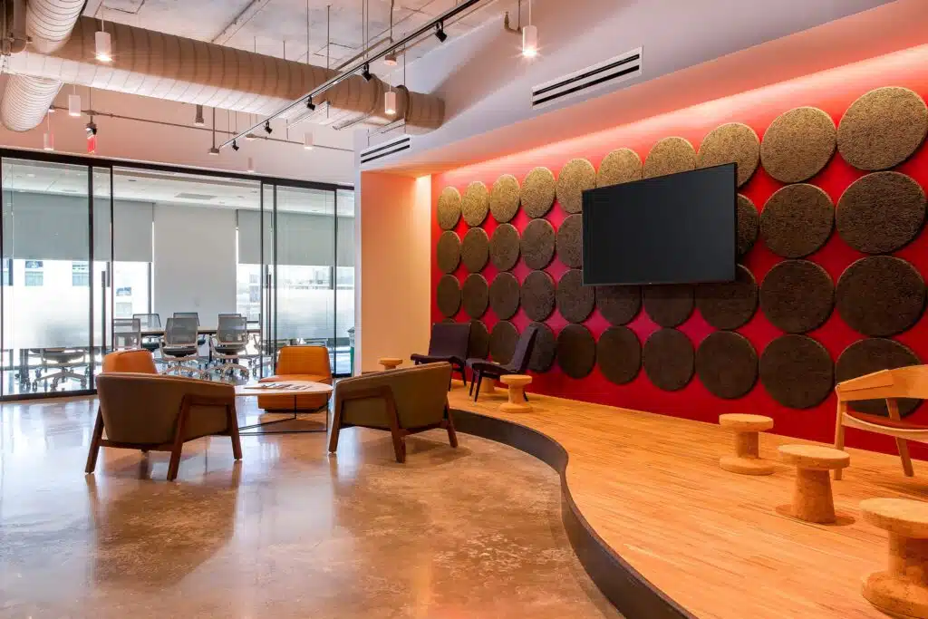 A modern office lounge area with a wall-mounted TV, circular wall decorations, comfortable chairs, small tables, and a glass-walled conference room in the background.