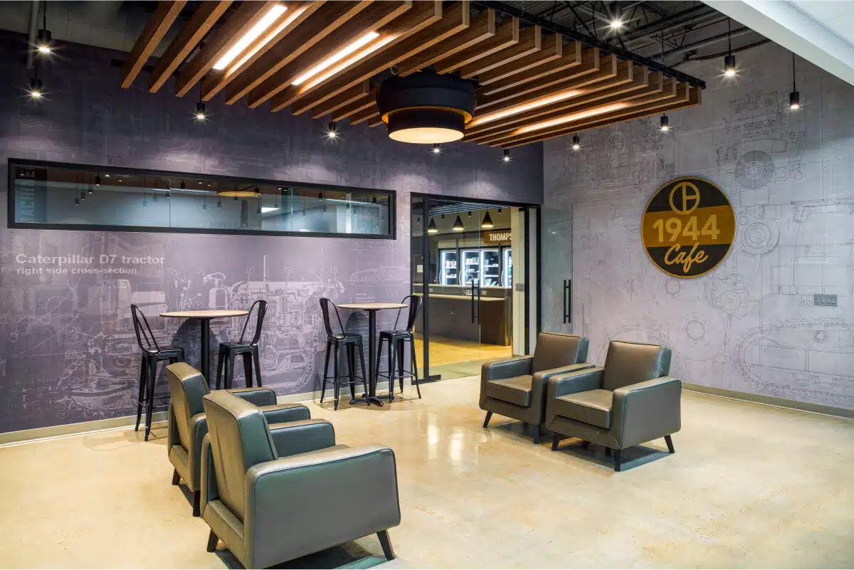 A modern café area featuring a few armchairs, high tables with stools, and a wall display with text and a logo reading "1944 Café." The room has industrial-style decor and a wooden ceiling feature.