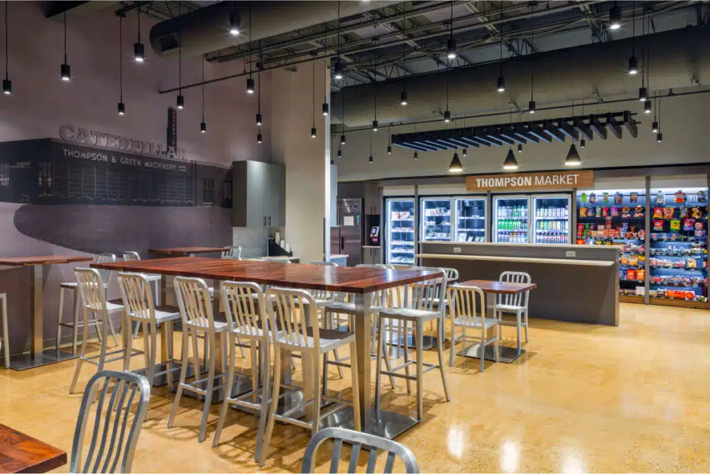 An indoor market with wooden tables and metal chairs, a counter labeled "Thompson Market," and shelves stocked with various beverages and snacks. The ceiling is decorated with hanging lights.