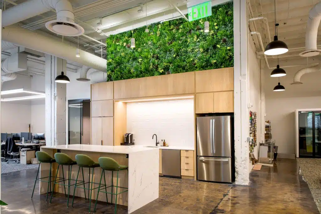 Modern office kitchen area with a large white marble island, green bar stools, stainless steel appliances, and a wall covered in greenery. Open office space is visible in the background.