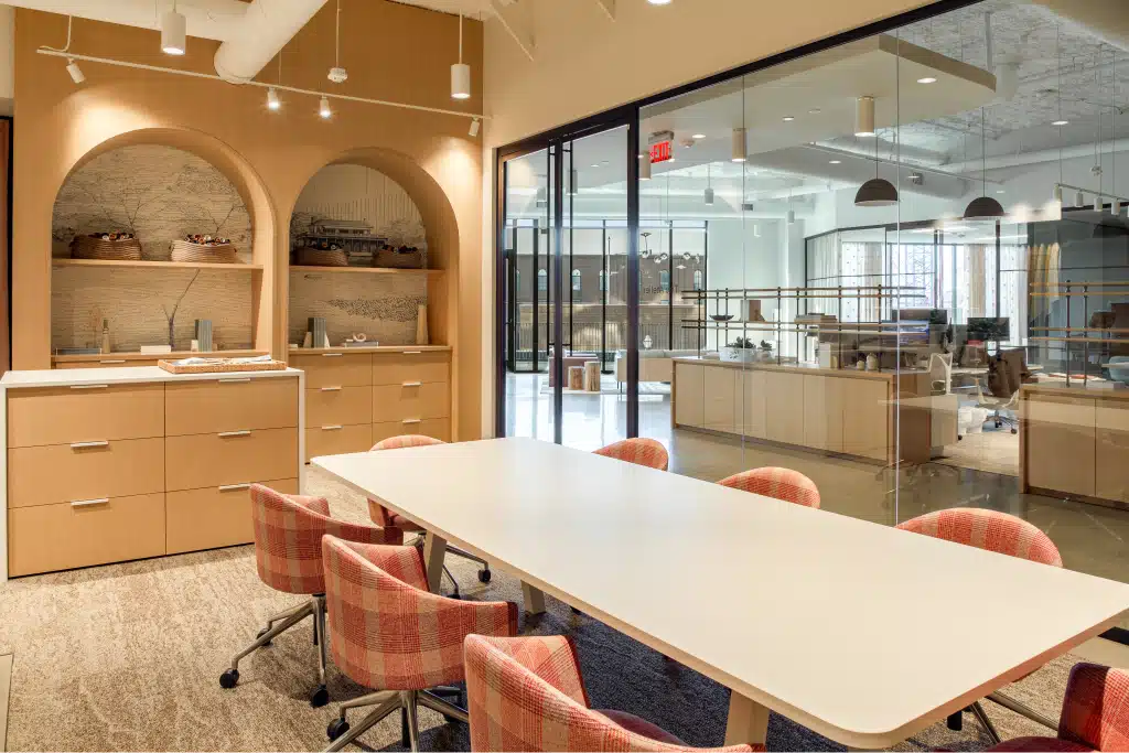 Modern conference room with a long white table, six plaid chairs, wood cabinetry, and glass wall partition revealing another office space.